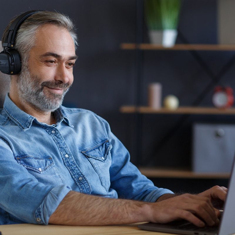 Portrait of handsome grey-haired man wearing headphones sitting at workplace. Senior male freelancer working in home office with laptop. Studying online, online courses.